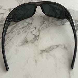May include: Dark grey sunglasses with a sporty design. The wraparound style sunglasses have dark lenses and a thick frame. The arms curve back for a secure fit. The lenses appear to be tinted.