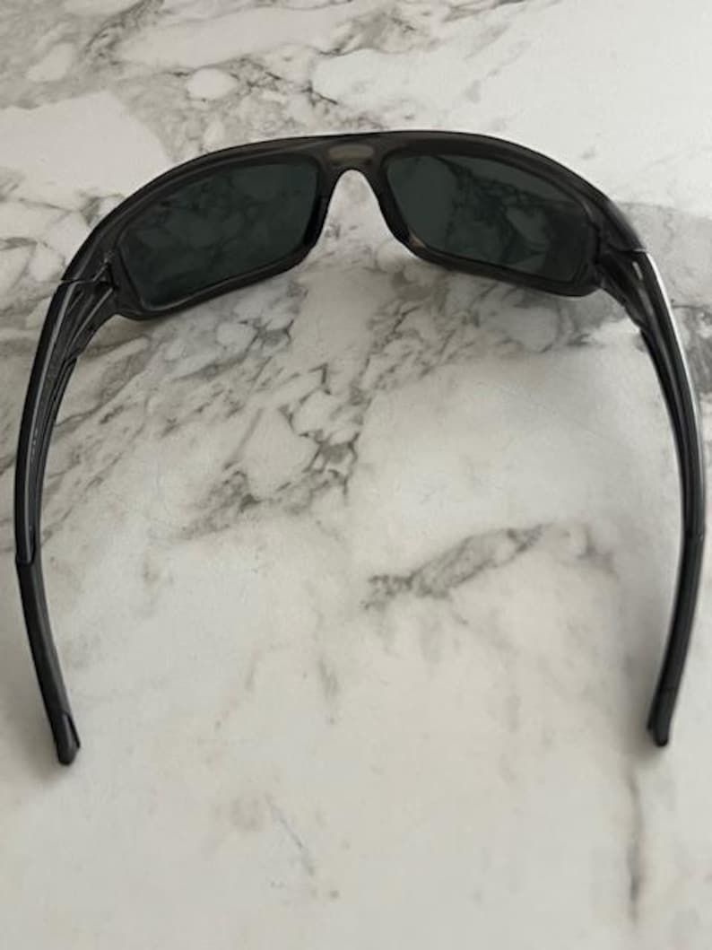 May include: Dark grey sunglasses with a sporty design. The wraparound style sunglasses have dark lenses and a thick frame. The arms curve back for a secure fit. The lenses appear to be tinted.