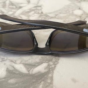May include: Dark gray sunglasses with a wraparound design. The lenses are a dark tint, and the frames have a subtle texture. The temples feature a light blue accent. These sunglasses are designed for outdoor activities.