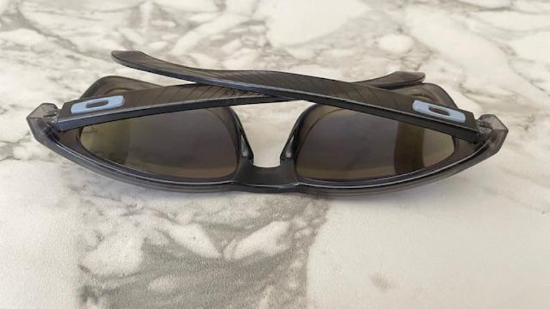 May include: Dark gray sunglasses with a wraparound design. The lenses are a dark tint, and the frames have a subtle texture. The temples feature a light blue accent. These sunglasses are designed for outdoor activities.