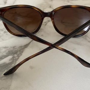 May include: Brown tortoiseshell sunglasses with dark lenses. The sunglasses have a classic, rounded frame shape and are viewed from the back, with the arms crossed. The lenses are a gradient brown color.