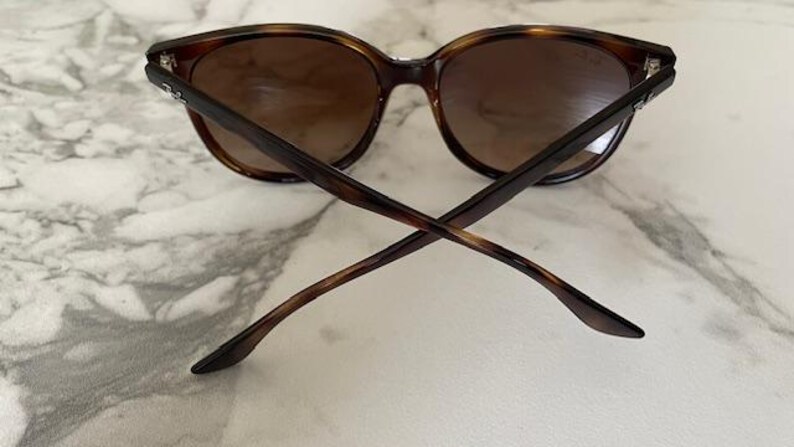 May include: Brown tortoiseshell sunglasses with dark lenses. The sunglasses have a classic, rounded frame shape and are viewed from the back, with the arms crossed. The lenses are a gradient brown color.