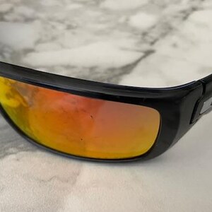 Puede incluir: Gafas de sol negras con lentes espejadas naranjas y amarillas. Las gafas tienen un dise&ntilde;o elegante y moderno con montura negra y logotipo plateado en las patillas. Las lentes reflejan un degradado de colores.