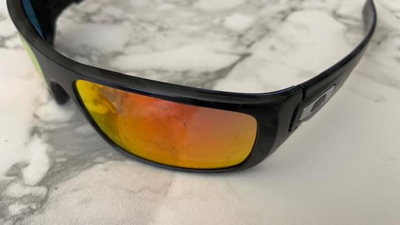Puede incluir: Gafas de sol negras con lentes espejadas naranjas y amarillas. Las gafas tienen un dise&ntilde;o elegante y moderno con montura negra y logotipo plateado en las patillas. Las lentes reflejan un degradado de colores.