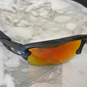 Peut inclure: Lunettes de soleil noires avec verres orange-rouge. Les lunettes ont un design &eacute;l&eacute;gant et moderne avec une monture et des branches noires. Les verres sont d'une couleur orange-rouge vibrante, offrant un look audacieux. Les lunettes reposent sur une surface en marbre blanc et gris.