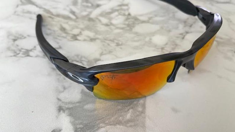 Peut inclure: Lunettes de soleil noires avec verres orange-rouge. Les lunettes ont un design &eacute;l&eacute;gant et moderne avec une monture et des branches noires. Les verres sont d'une couleur orange-rouge vibrante, offrant un look audacieux. Les lunettes reposent sur une surface en marbre blanc et gris.