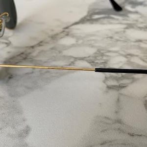 May include: Gold-rimmed aviator sunglasses with dark lenses. The sunglasses have a classic teardrop shape and a thin gold frame. The temples are gold with black earpieces. The sunglasses are resting on a marble surface.