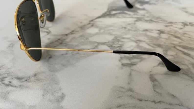 May include: Gold-rimmed aviator sunglasses with dark lenses. The sunglasses have a classic teardrop shape and a thin gold frame. The temples are gold with black earpieces. The sunglasses are resting on a marble surface.