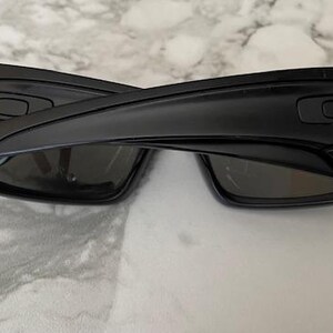 May include: Black sunglasses featuring a gold anchor emblem on each arm. The sunglasses have a rectangular frame and dark lenses. The arms are thick and curve around the ears. The sunglasses are resting on a marble surface.