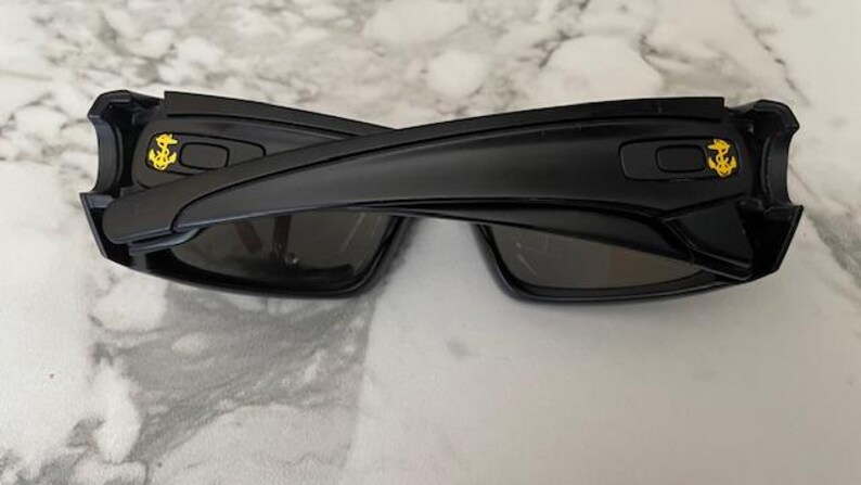 May include: Black sunglasses featuring a gold anchor emblem on each arm. The sunglasses have a rectangular frame and dark lenses. The arms are thick and curve around the ears. The sunglasses are resting on a marble surface.