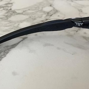 Puede incluir: Primer plano de gafas de sol negras con una patilla curva y una lente tintada. La patilla tiene un dise&ntilde;o texturizado y la marca es visible. Las gafas de sol est&aacute;n sobre una superficie similar al m&aacute;rmol.