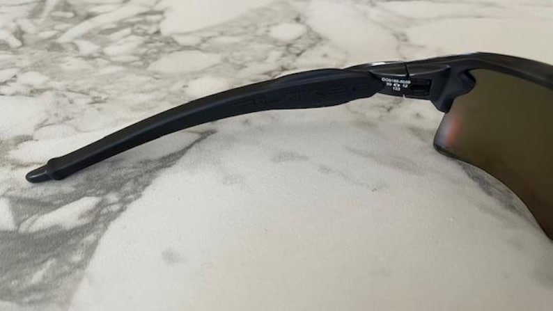 Puede incluir: Primer plano de gafas de sol negras con una patilla curva y una lente tintada. La patilla tiene un dise&ntilde;o texturizado y la marca es visible. Las gafas de sol est&aacute;n sobre una superficie similar al m&aacute;rmol.