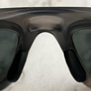 May include: Close-up of black sunglasses with a dark grey frame. The sunglasses have a curved bridge and black nose pads. The lenses are a dark grey colour.