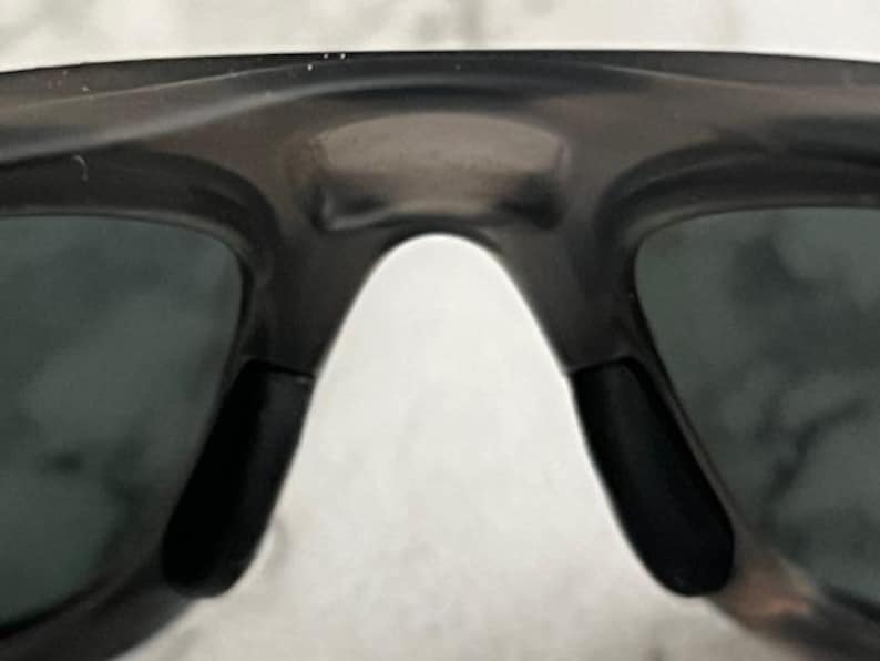 May include: Close-up of black sunglasses with a dark grey frame. The sunglasses have a curved bridge and black nose pads. The lenses are a dark grey colour.