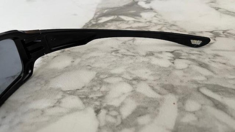 K&ouml;nnte beinhalten: Schwarze Sonnenbrille mit rechteckiger Linsenform. Der Rahmen ist schwarz und hat ein schlankes, modernes Design. Der B&uuml;gel hat ein kleines, geometrisches Detail am Ende.