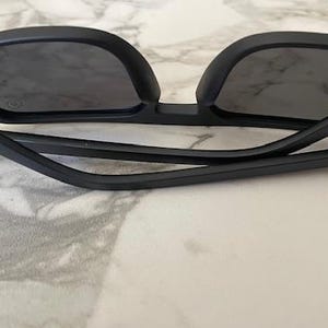 Puede incluir: Gafas de sol negro mate con lentes oscuras y montura plegada. Las patillas presentan un sutil detalle de dos l&iacute;neas cerca de las bisagras. Las gafas est&aacute;n colocadas con las patillas plegadas hacia adentro, mostrando la forma rectangular.