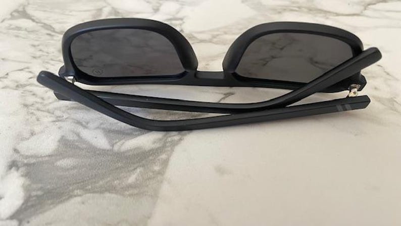 Puede incluir: Gafas de sol negro mate con lentes oscuras y montura plegada. Las patillas presentan un sutil detalle de dos l&iacute;neas cerca de las bisagras. Las gafas est&aacute;n colocadas con las patillas plegadas hacia adentro, mostrando la forma rectangular.