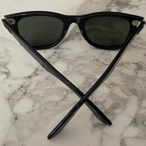 K&ouml;nnte beinhalten: Schwarze Sonnenbrille mit dunklen Gl&auml;sern. Die Sonnenbrille hat ein klassisches, eckiges Rahmendesign. Die B&uuml;gel sind gekreuzt, und die Brille liegt auf einem Marmorhintergrund. Ein stilvolles Accessoire zum Schutz der Augen.