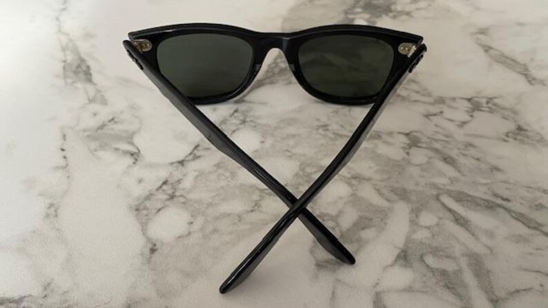 K&ouml;nnte beinhalten: Schwarze Sonnenbrille mit dunklen Gl&auml;sern. Die Sonnenbrille hat ein klassisches, eckiges Rahmendesign. Die B&uuml;gel sind gekreuzt, und die Brille liegt auf einem Marmorhintergrund. Ein stilvolles Accessoire zum Schutz der Augen.