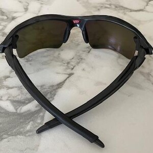 Puede incluir: Gafas de sol negras con lentes oscuras, vistas desde atr&aacute;s, sobre una superficie de m&aacute;rmol. Las patillas de las gafas est&aacute;n cruzadas. La parte superior de la montura tiene la palabra "USA" en rojo.