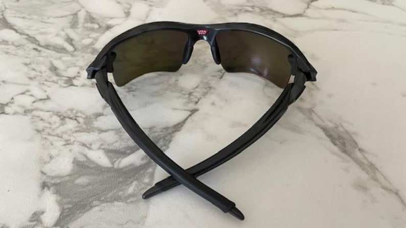 Puede incluir: Gafas de sol negras con lentes oscuras, vistas desde atr&aacute;s, sobre una superficie de m&aacute;rmol. Las patillas de las gafas est&aacute;n cruzadas. La parte superior de la montura tiene la palabra "USA" en rojo.