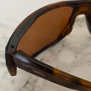 Op de afbeelding: Close-up van een Oakley zonnebril met een montuur met schildpadpatroon en bruine glazen. De merknaam "OAKLEY" is zichtbaar op de poot. De zonnebril is ontworpen voor buitenactiviteiten en biedt bescherming tegen de zon.