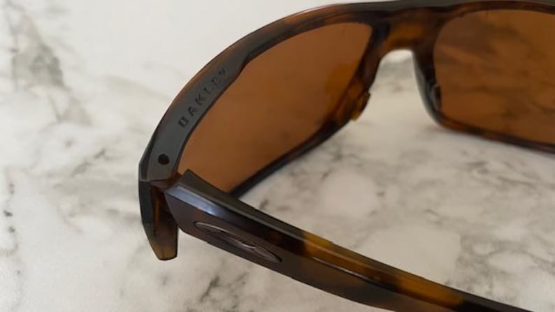 Op de afbeelding: Close-up van een Oakley zonnebril met een montuur met schildpadpatroon en bruine glazen. De merknaam "OAKLEY" is zichtbaar op de poot. De zonnebril is ontworpen voor buitenactiviteiten en biedt bescherming tegen de zon.