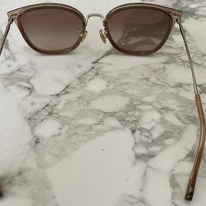May include: Cat-eye sunglasses with a gradient brown lens and a gold-tone metal frame. The temples are a brown color. The sunglasses are resting on a white and gray marble surface.
