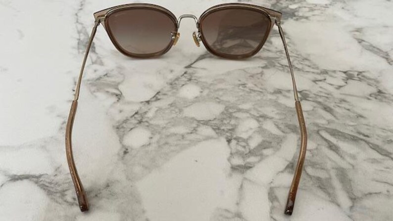 May include: Cat-eye sunglasses with a gradient brown lens and a gold-tone metal frame. The temples are a brown color. The sunglasses are resting on a white and gray marble surface.