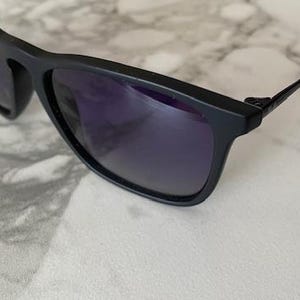 May include: Black sunglasses with dark purple lenses. The sunglasses have a rectangular frame and thin, black arms. The lenses reflect the surrounding environment. The sunglasses are resting on a white and gray marble surface.