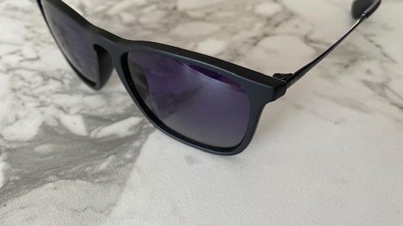 May include: Black sunglasses with dark purple lenses. The sunglasses have a rectangular frame and thin, black arms. The lenses reflect the surrounding environment. The sunglasses are resting on a white and gray marble surface.