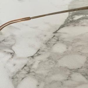 May include: Close-up of a pair of sunglasses with a gold-colored frame and brown lenses. The temple arm is a thin, gold metal, and the earpiece is a translucent, light brown plastic. The sunglasses rest on a white and gray marble surface.