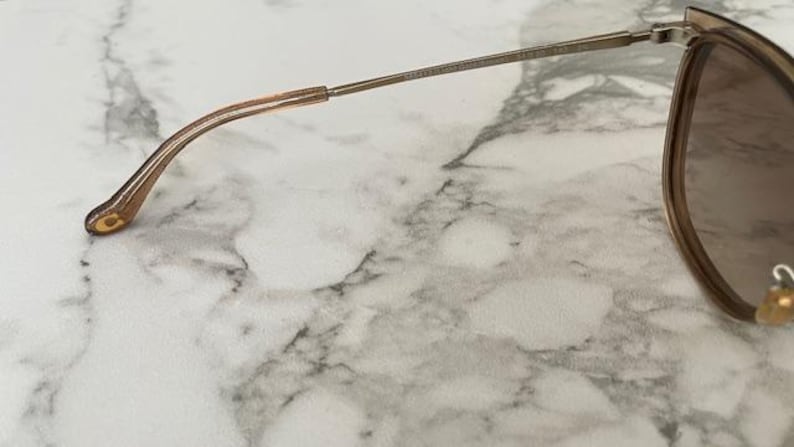 May include: Close-up of a pair of sunglasses with a gold-colored frame and brown lenses. The temple arm is a thin, gold metal, and the earpiece is a translucent, light brown plastic. The sunglasses rest on a white and gray marble surface.