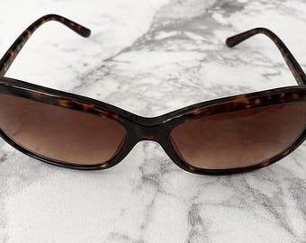 Puede incluir: Gafas de sol carey con lentes degradadas marrones. Las monturas tienen una forma clásica, ligeramente ojo de gato. Las gafas de sol están sobre un fondo de mármol.