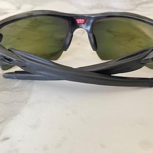 Puede incluir: Gafas de sol deportivas negras con lentes de color verde. Las gafas de sol tienen un dise&ntilde;o elegante y moderno con montura y patillas negras. El puente de las gafas tiene el texto "USA" en rojo. Las patillas est&aacute;n plegadas.