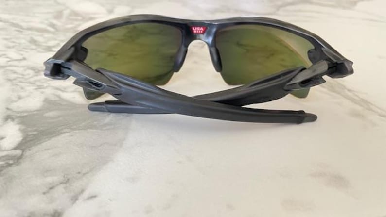 Puede incluir: Gafas de sol deportivas negras con lentes de color verde. Las gafas de sol tienen un dise&ntilde;o elegante y moderno con montura y patillas negras. El puente de las gafas tiene el texto "USA" en rojo. Las patillas est&aacute;n plegadas.