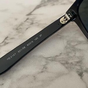 Puede incluir: Primer plano de gafas de sol negras con lentes oscuras. La patilla de las gafas de sol muestra los n&uacute;meros "RB4147 601/58 60015 145 3P". Las gafas de sol est&aacute;n sobre un fondo de m&aacute;rmol.