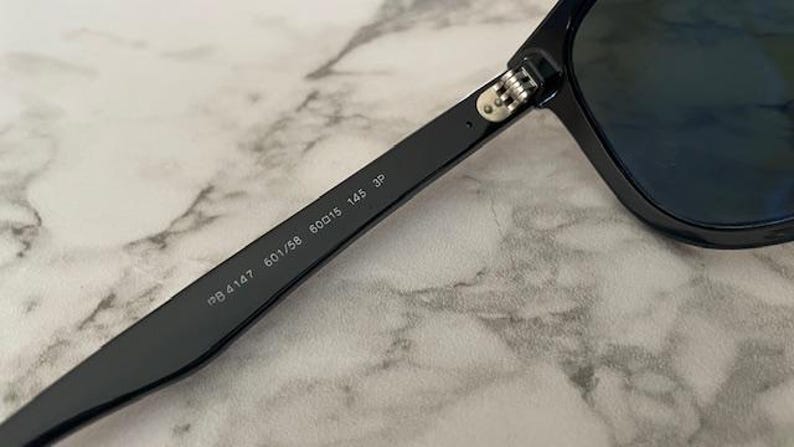Puede incluir: Primer plano de gafas de sol negras con lentes oscuras. La patilla de las gafas de sol muestra los n&uacute;meros "RB4147 601/58 60015 145 3P". Las gafas de sol est&aacute;n sobre un fondo de m&aacute;rmol.