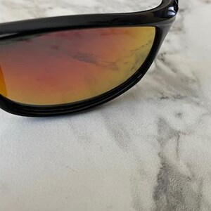 May include: Close-up of black sunglasses with orange and red gradient lenses. The sunglasses have a sleek, curved design and are set against a white and gray marble surface. The lenses reflect light, creating a vibrant color gradient.