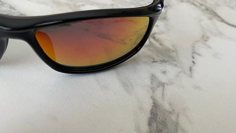 May include: Close-up of black sunglasses with orange and red gradient lenses. The sunglasses have a sleek, curved design and are set against a white and gray marble surface. The lenses reflect light, creating a vibrant color gradient.