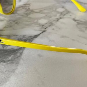May include: Bright yellow sunglasses with a classic frame design. The Ray-Ban logo is visible on the temple. The sunglasses are set against a marble-patterned surface.