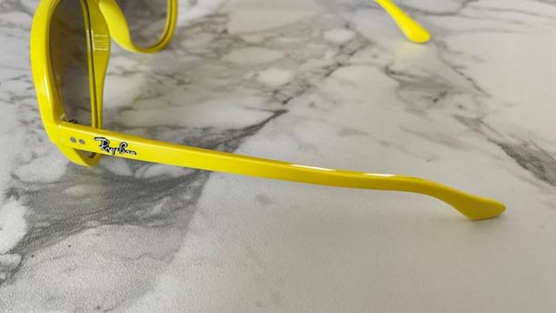 May include: Bright yellow sunglasses with a classic frame design. The Ray-Ban logo is visible on the temple. The sunglasses are set against a marble-patterned surface.