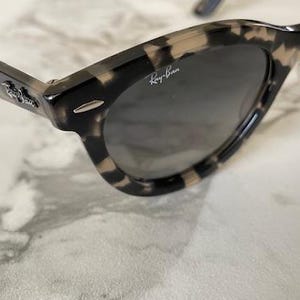Puede incluir: Gafas de sol con estampado de carey y lentes oscuras. Las gafas de sol tienen un dise&ntilde;o cl&aacute;sico con una montura gruesa y el logotipo de Ray-Ban visible en la lente. Las patillas tambi&eacute;n tienen un estampado.