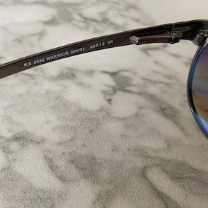 May include: Close-up of a pair of sunglasses with a dark metal frame and gradient lenses. The text "RB 3342 WARRIOR 004/51 60[]12 2N" is printed on the temple arm. The lenses have a brown tint.