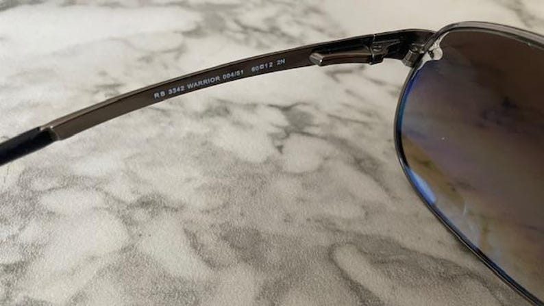 May include: Close-up of a pair of sunglasses with a dark metal frame and gradient lenses. The text "RB 3342 WARRIOR 004/51 60[]12 2N" is printed on the temple arm. The lenses have a brown tint.