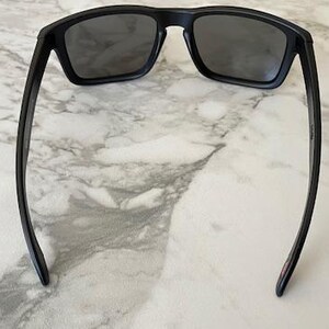 May include: Black sunglasses with a rectangular frame and dark lenses. The sunglasses have a sleek, modern design with straight arms. The image is taken from a top-down perspective, showcasing the sunglasses against a marble surface.
