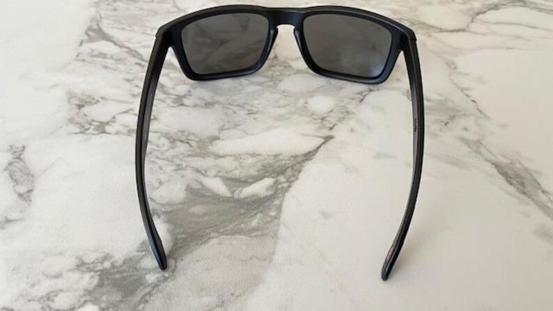 May include: Black sunglasses with a rectangular frame and dark lenses. The sunglasses have a sleek, modern design with straight arms. The image is taken from a top-down perspective, showcasing the sunglasses against a marble surface.
