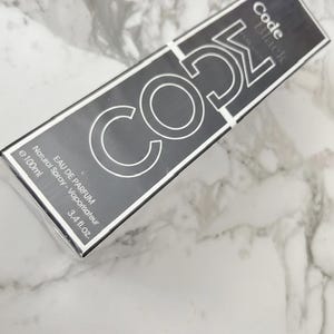 May include: A rectangular black perfume box with the text "Code Black" and "CO52" in a silver, bold font. The box also includes the text "EAU DE PARFUM" and "3.4 fl.oz.". The box is set against a marble background.
