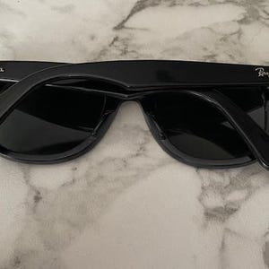 K&ouml;nnte beinhalten: Schwarze Sonnenbrille mit dunklen Gl&auml;sern, mit dem Ray-Ban-Logo auf den B&uuml;geln. Die Sonnenbrille liegt auf einem Marmorhintergrund. Die Rahmen sind gl&auml;nzend und reflektieren das Licht.