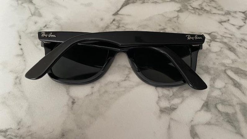 K&ouml;nnte beinhalten: Schwarze Sonnenbrille mit dunklen Gl&auml;sern, mit dem Ray-Ban-Logo auf den B&uuml;geln. Die Sonnenbrille liegt auf einem Marmorhintergrund. Die Rahmen sind gl&auml;nzend und reflektieren das Licht.